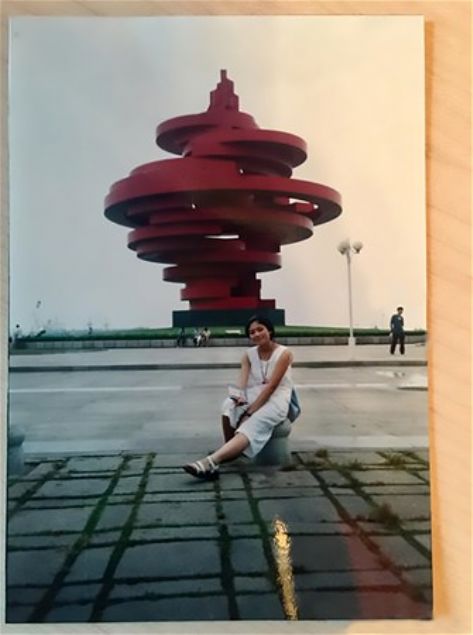  Xue, Beyaz elbisesiyle devasa krmz heykelin nnde poz verirken arkada gen ocuk ise gelecekteki kocas Ye.         